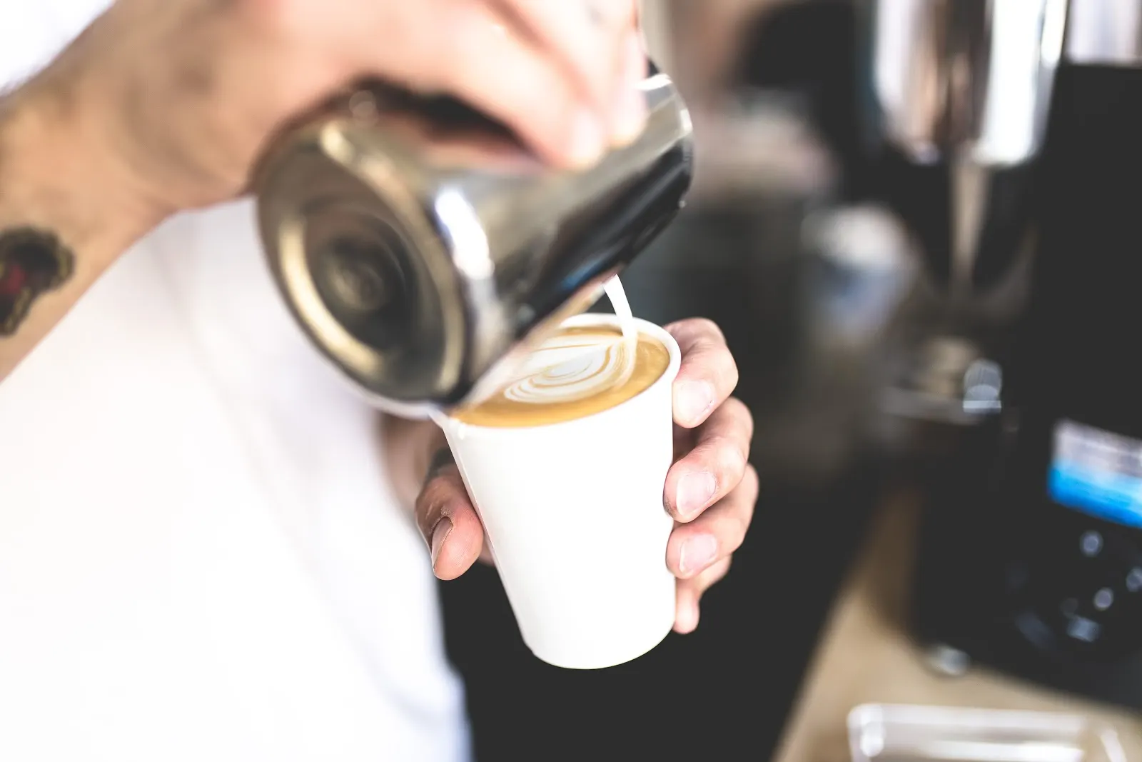 Barista pouring steamed milk from metal pitcher into white cup, creating coffee.