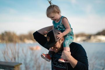 A person carrying a small child on their shoulders near a body of water at sunset.